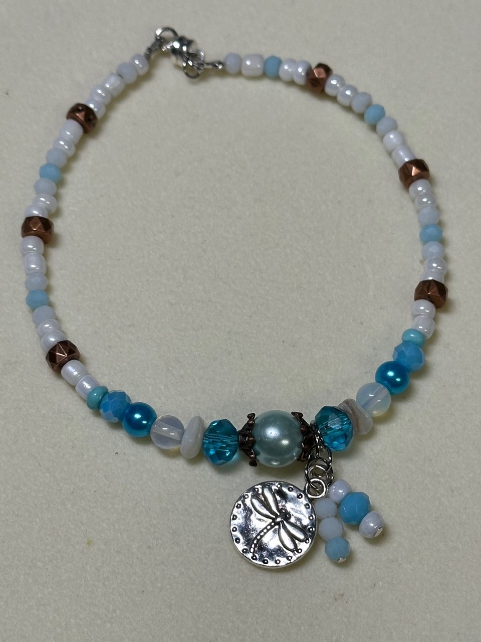 Anklet with a variety of different colored beads. There is a small charm in the middle of a dragonfly